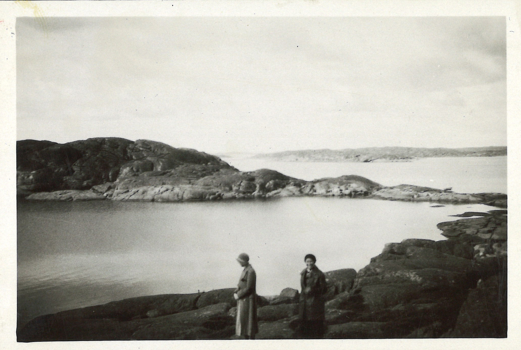 
                    Ett svartvitt fotografi av två kvinnor i Göteborgs skärgård. De har kappor och hatt på sig. En av dem tittar mot kameran och en tittar ut mot havet. 
                    