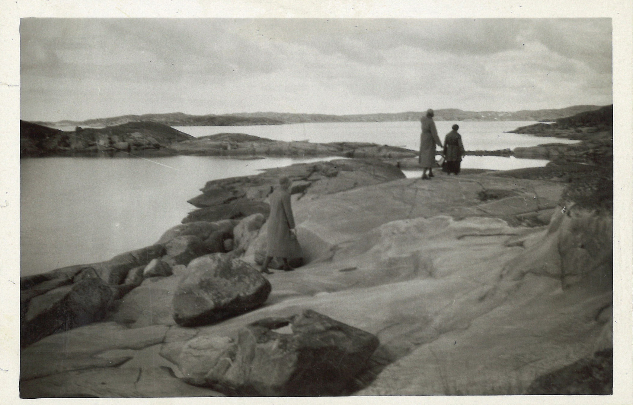 
                    Ett svartvitt fotografi av tre kvinnor som promenerar över klipporna i Göteborgs skärgård. De har alla tre ryggarna mot kameran. 
                    
