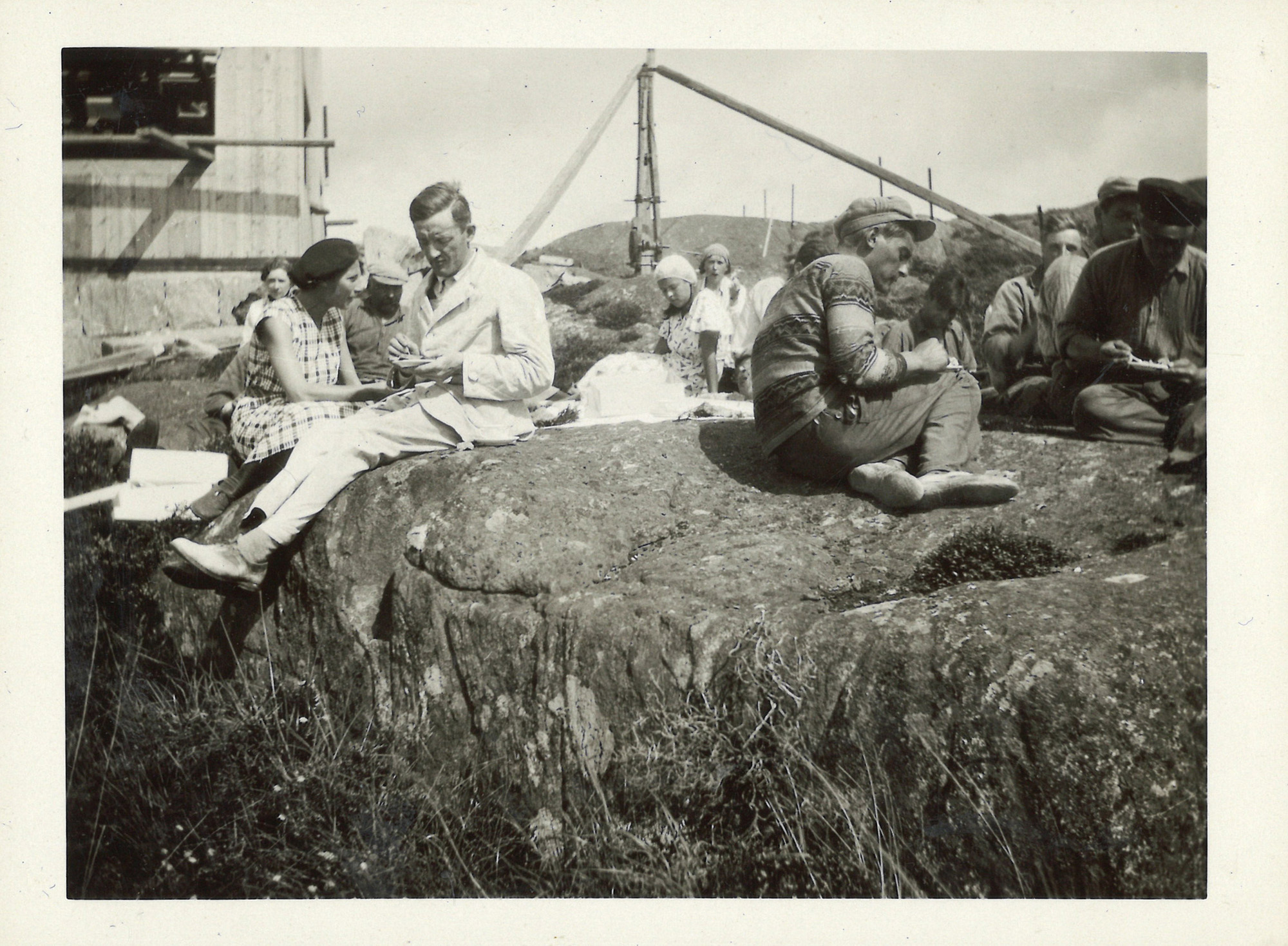 
                    Ett svartvitt fotografi av män och kvinnor som sitter på några klippor och fikar. I bakgrunden skymtar fasaden på systrarnas sommarstuga. Den är omålad och fönstren har inte än blivit insatta. 
                    