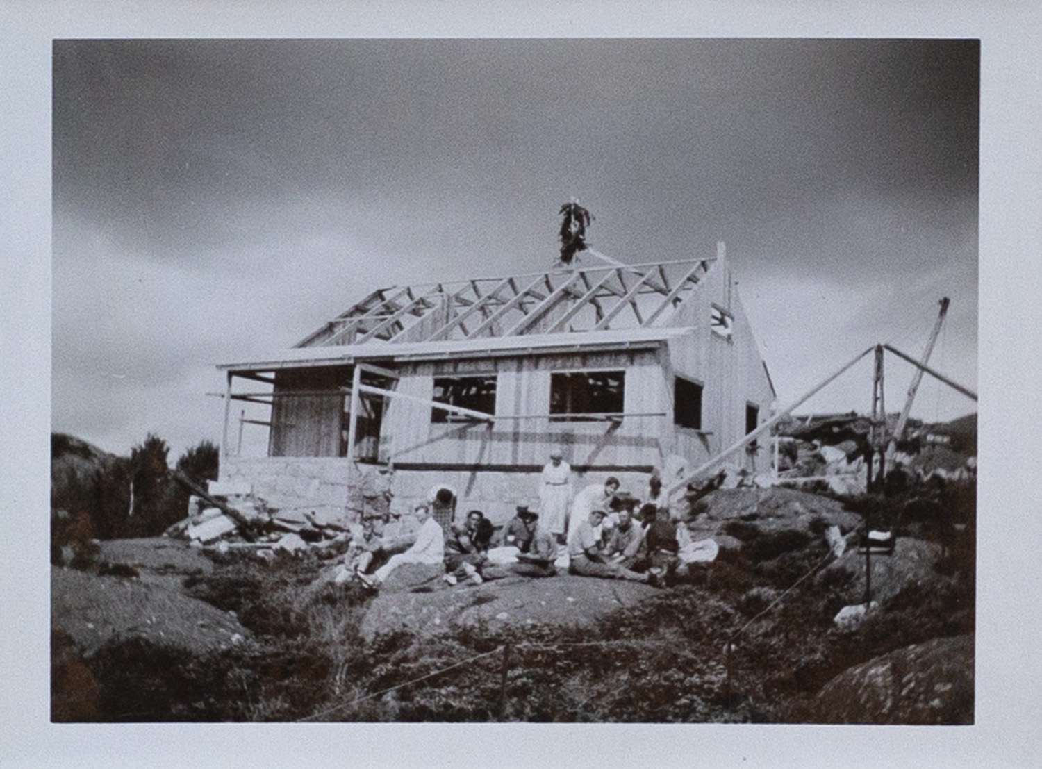 
                    Ett svartvitt fotografi av stugan under byggnation. Fasaden är rest men fönster och tak saknas. På en påle som sticker upp över taknocken är ett ris fastsatt. Framför byggnaden sitter en samling män och kvinnor på klipporna.  
                    