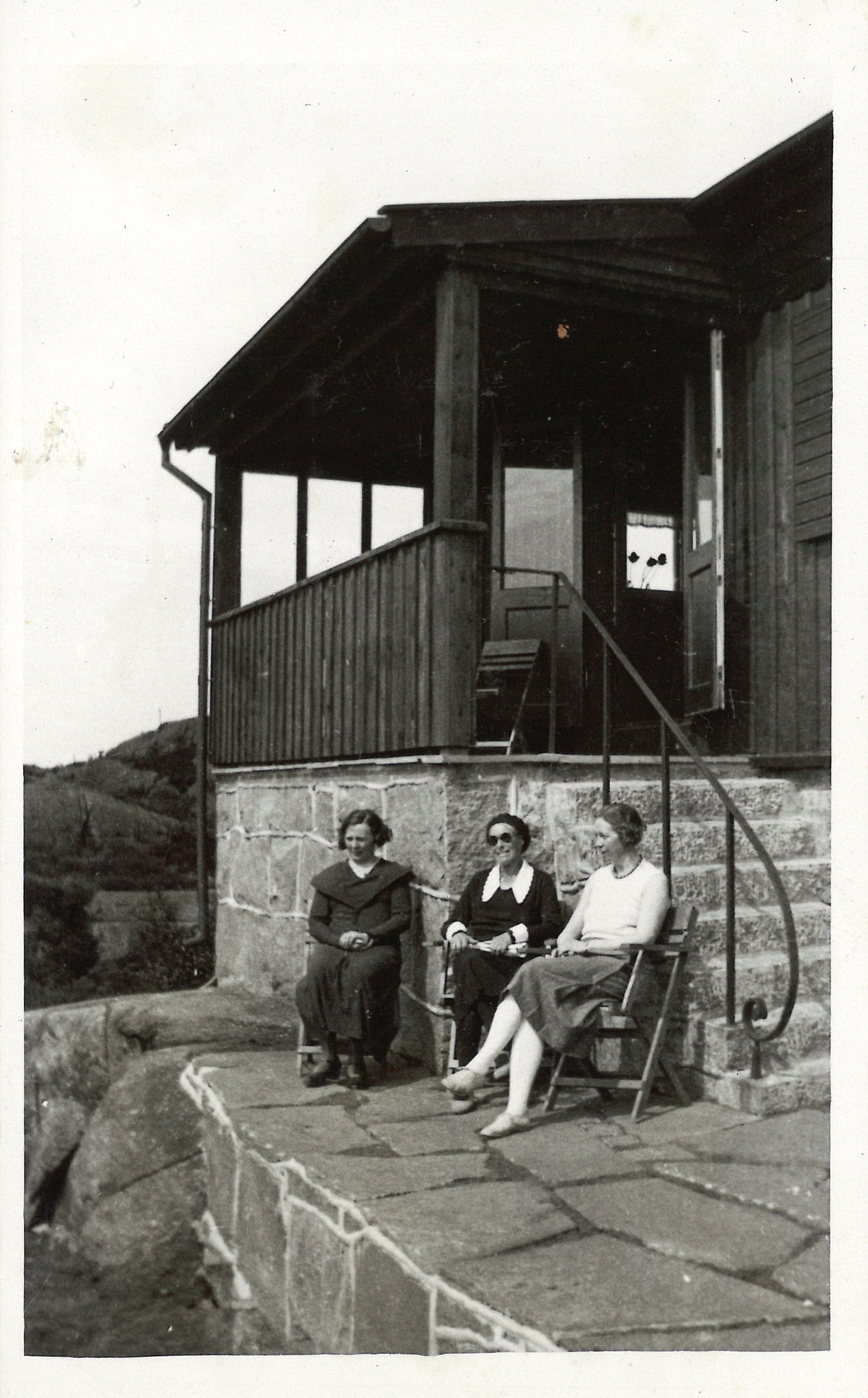
                    Ett svartvitt fotografi av tre kvinnor som sitter på stolar framför sommarstugan. Bakom den syns stugans trappa som går upp till en veranda, där dörrarna in till stugan står öppna.  
                    