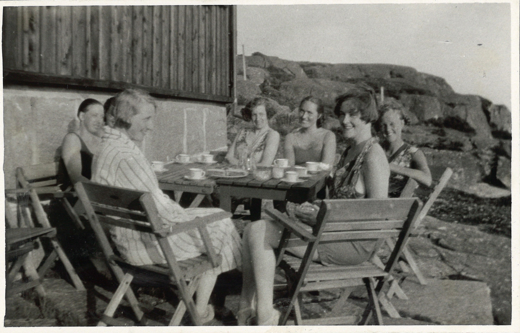 
                    Ett svartvitt fotografi av sju kvinnor som sitter runt ett bord utanför sommarstugan. Flera av kvinnorna har badkläder på sig och på bordet står det kaffekoppar och en kaka.  
                    
